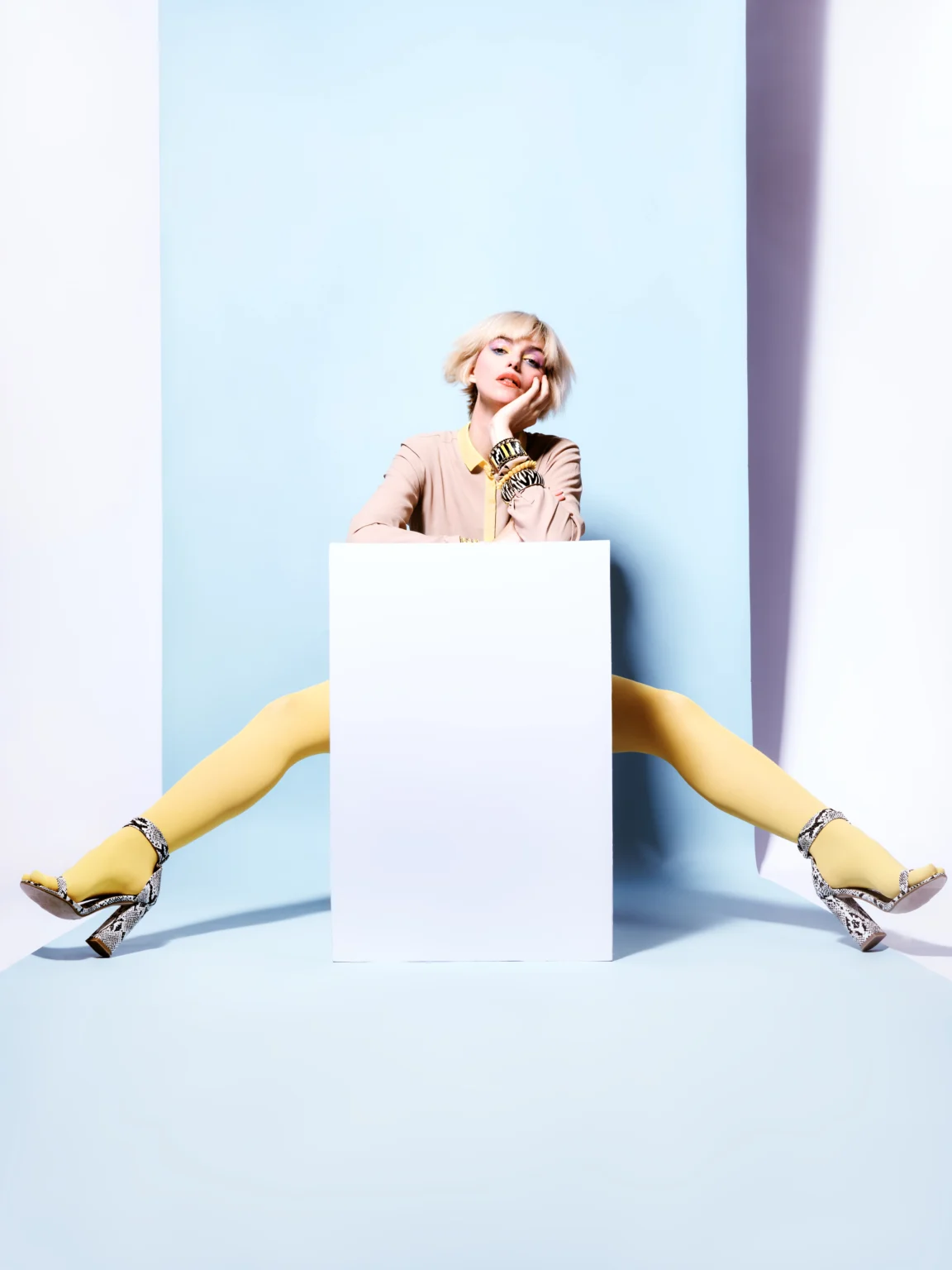 Femme assise derrière un cube blanc, les jambes écartées en pantalon jaune.